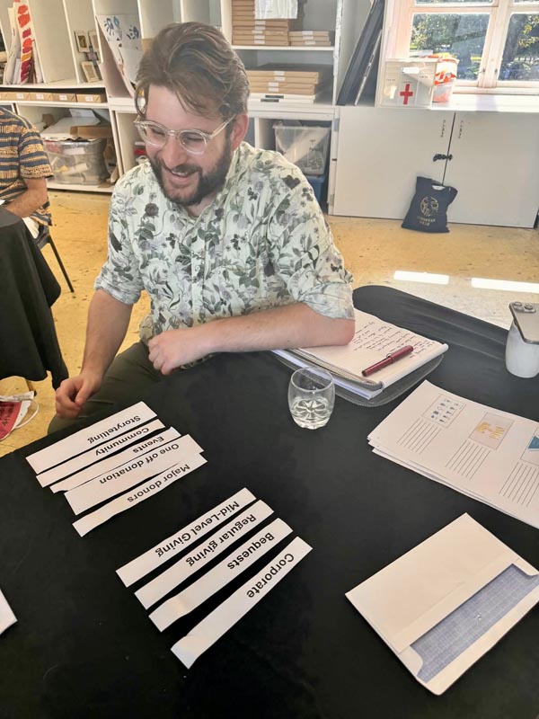 Caleb Gordon is seated at a table with  a dozen strips of paper with words like "corportate", "bequests" and "regular giving" on them